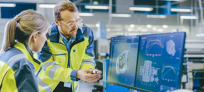 Two people wearing high‑visibility work jackets stand in an industrial or manufacturing environment, reviewing detailed technical diagrams, charts, and performance graphs displayed on multiple computer monitors.